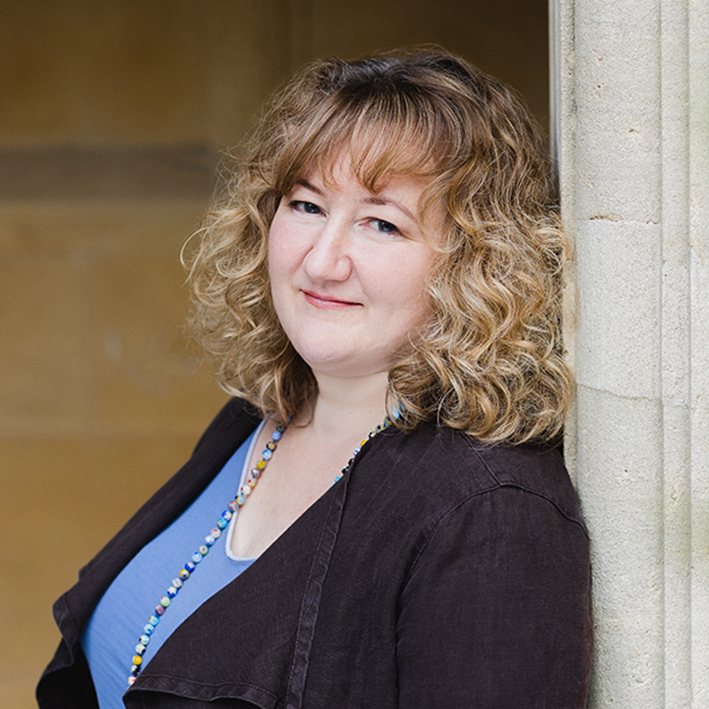 Woman with curly blonde hair and blue eyes smiles at the camera as she leans against a stone pillar. She is wearing a brown jacket over a Madonna blue shirt and a long beaded necklace.